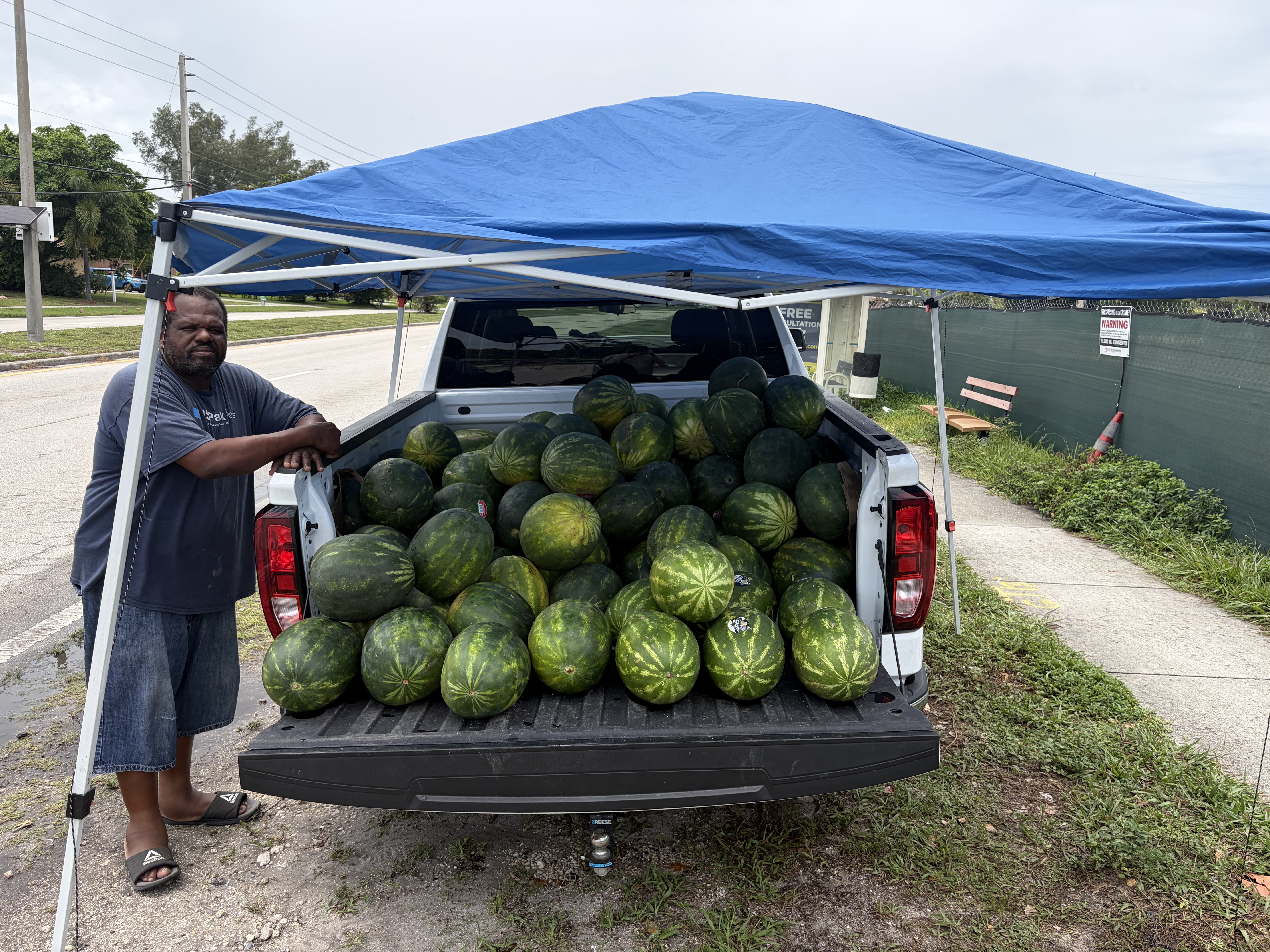 Derek beside his pickup truck stacked with watermelons under a blue canopy, roadside on Australian Avenue